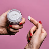 Sur un fond rose, l'index de la main droite a été plongé dans un pot d'huile de massage comestible pour montrer la texture crémeuse du produit