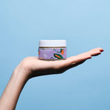 Au creux d'une main sur un fond bleu ciel, une pot d'huile de massage en verre de la marque Goliate