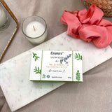 Un étui carton de savon solide Cosmoz anti-imperfections posé sur une plaque de marbre avec un chouchou satiné rose et une bougie blanche