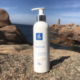 Dans un décor de plage bretonne rocailleuse, le lait corps aux huiles essentielles d'agrumes est posé sur un rocher. En arrière-plan on aperçoit la mer
