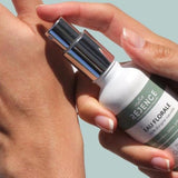 Sur fond bleu clair, une main de femme tient un flacon d'eau florale Réjence pour le cuir chevelu