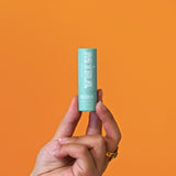 Sur un fond orange, une main de femme portant une bague dorée tient entre ses doigts un baume à lèvres de la marque My Mira