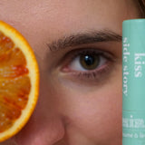 Gros plan sur le visage d'une femme blanche, au premier plan on voit un tiers d'une tranche d'orange et un tube de baume à lèvres Kiss Side Story de la marque My Mira de couleur bleu-vert
