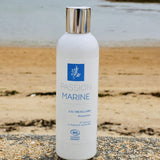 Un flacon d'eau micellaire démaquillante Passion Marine est posé sur un mur et en arrière-plan on voit une plage de sable en Bretagne avec l'océan et quelques algues