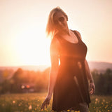 Une femme blanche, blonde, avec des lunettes de soleil teintées noires, porte une robe noire au-dessus du genou dans un champ de fleurs jaunes au coucher du soleil