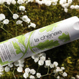 Un flacon de lotion la Chênaie posé sur de la mousse et entourée de branches de petites fleurs blanches