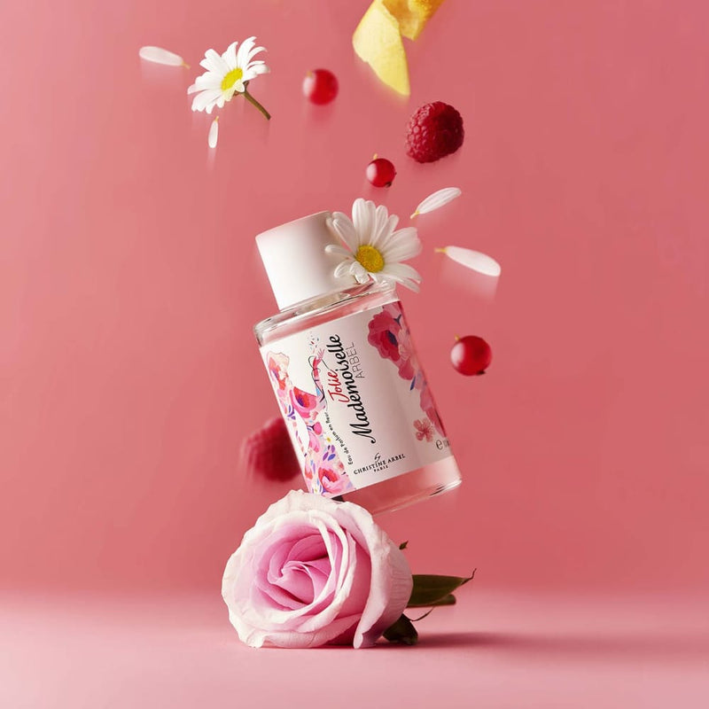 Sur un fond rose, un flacon de l'eau de parfum Jolie Mademoiselle Arbel avec en pied une rose de couleur rose pâle et au-dessus une envolée de groseilles, framboises, marguerites et zeste d'agrumes
