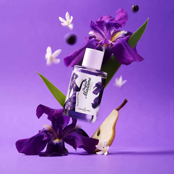 Sur un fond violet, un flacon de l'eau de parfum Madame Arbel avec deux iris violettes, quelques cassis et des fleurs blanches