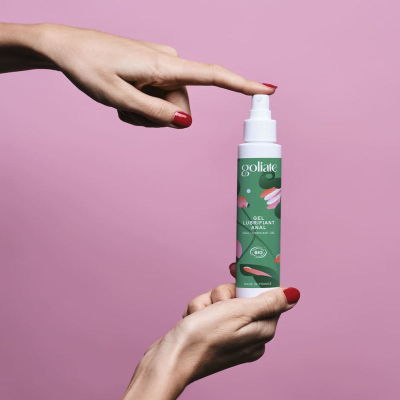 Sur fond rose, des mains de femme tiennent un flacon de gel lubrifiant anal de la marque Goliate