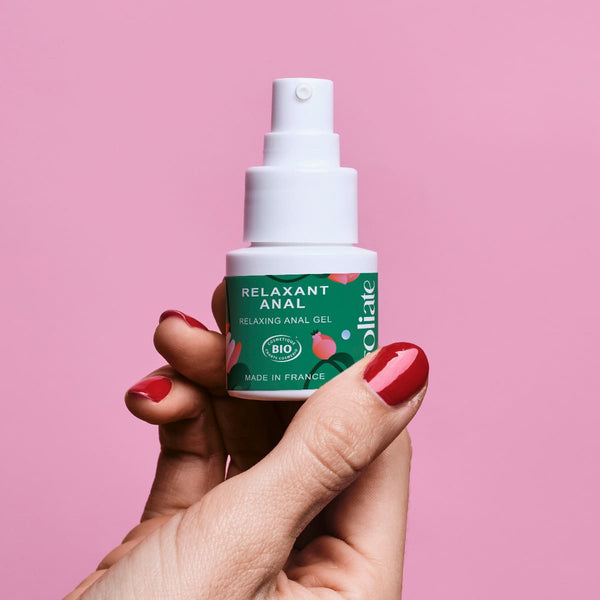 Sur un fond rose, une main de femme avec du vernis à ongles rouge tient un flacon de gel relaxant anal de la marque Goliate