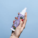 Sur un fond bleu clair, une main de femme tient un flacon d'huile de massage aphrodisiaque de la marque Goliate