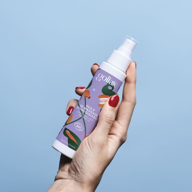 Sur un fond bleu clair, une main de femme tient un flacon d'huile de massage aphrodisiaque de la marque Goliate