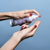 Sur un fond bleu, on voit les mains vernies de rouge d'une femme tenir un flacon d'huile de massage aphrodisiaque de la marque Goliate. Elle appuie sur la pompe pour montrer la texture de l'huile sur son index droit.