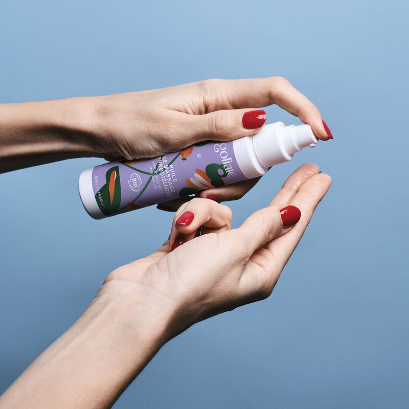 Sur un fond bleu, on voit les mains vernies de rouge d'une femme tenir un flacon d'huile de massage aphrodisiaque de la marque Goliate. Elle appuie sur la pompe pour montrer la texture de l'huile sur son index droit.