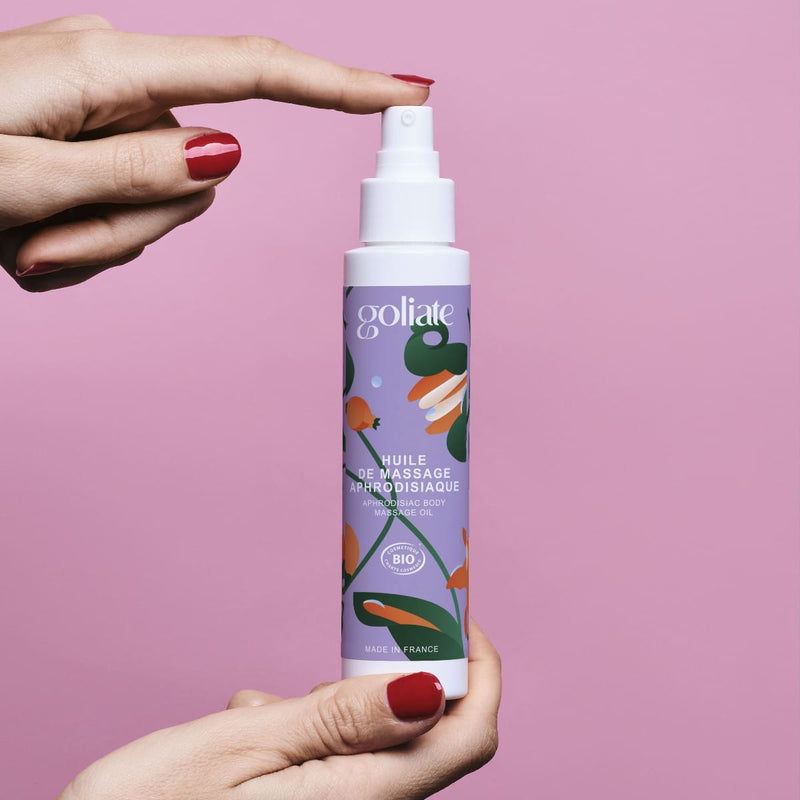 Sur un fond rose, des mains de femme tiennent un flacon d'huile de massage aphrodisiaque de la marque Goliate