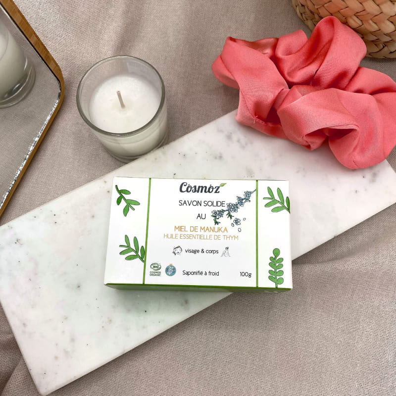 Un étui carton de savon solide Cosmoz anti-imperfections posé sur une plaque de marbre avec un chouchou satiné rose et une bougie blanche