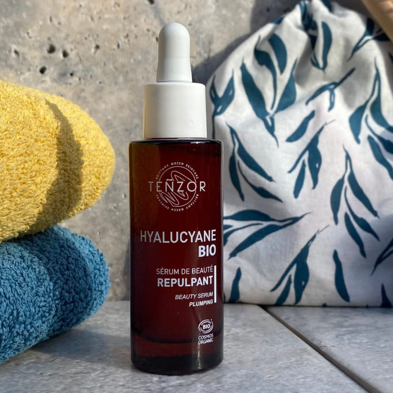 Sur du carrelage gris, un flacon-pipette de sérum repulpant Hyalucyane à l'acide hyaluronique avec deux serviettes de bain et un pochon en tissu