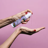 Sur un fond rose, des mains de femme tiennent un flacon d'huile de massage aphrodisiaque Goliate et montrent la texture