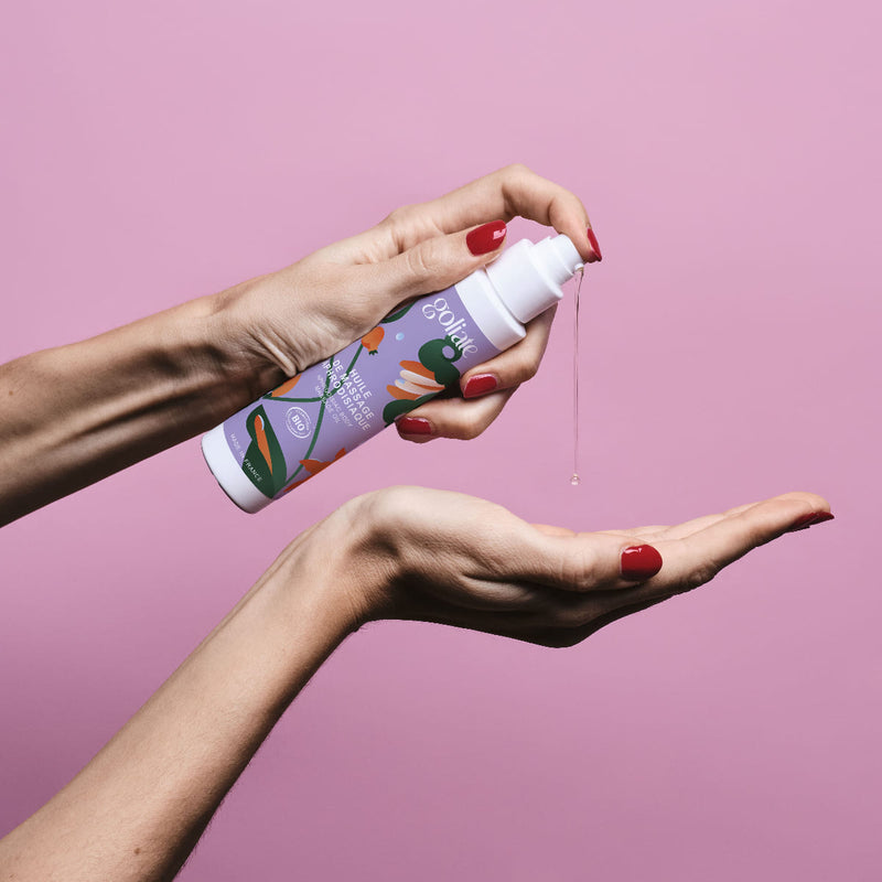 Sur un fond rose, des mains de femme tiennent un flacon d'huile de massage aphrodisiaque Goliate et montrent la texture