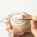 Une main de femme tient un pot en verre avec à l'intérieur le déodorant peaux sensibles de la marque Endro. Sur une spatule tenue au-dessus du pot, une noisette de déodorant est déposée pour montrer sa texture.