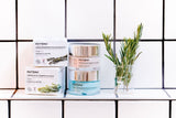 Dans une salle de bain avec du carrelage blanc, deux étuis cartons de crèmes pour le visage Phytema, à côté deux pots de crèmes et un récipient avec des branches de romarin