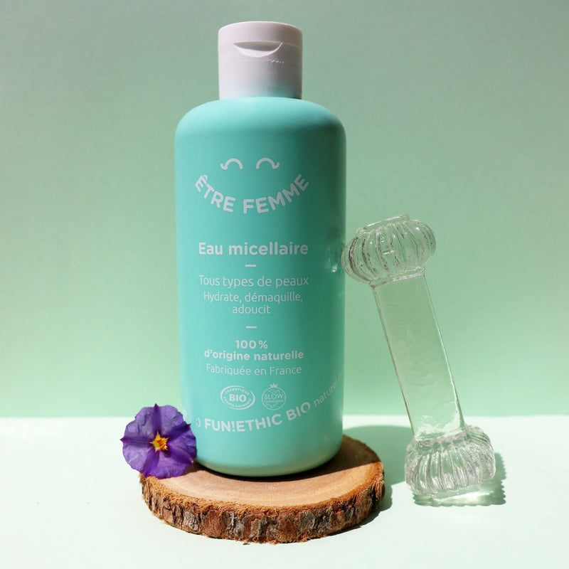 Une eau micellaire de la gamme Être Femme de Fun!Ethic est posée sur une coupe de bois avec une petite pensée violette à gauche sur un fond vert turquoise